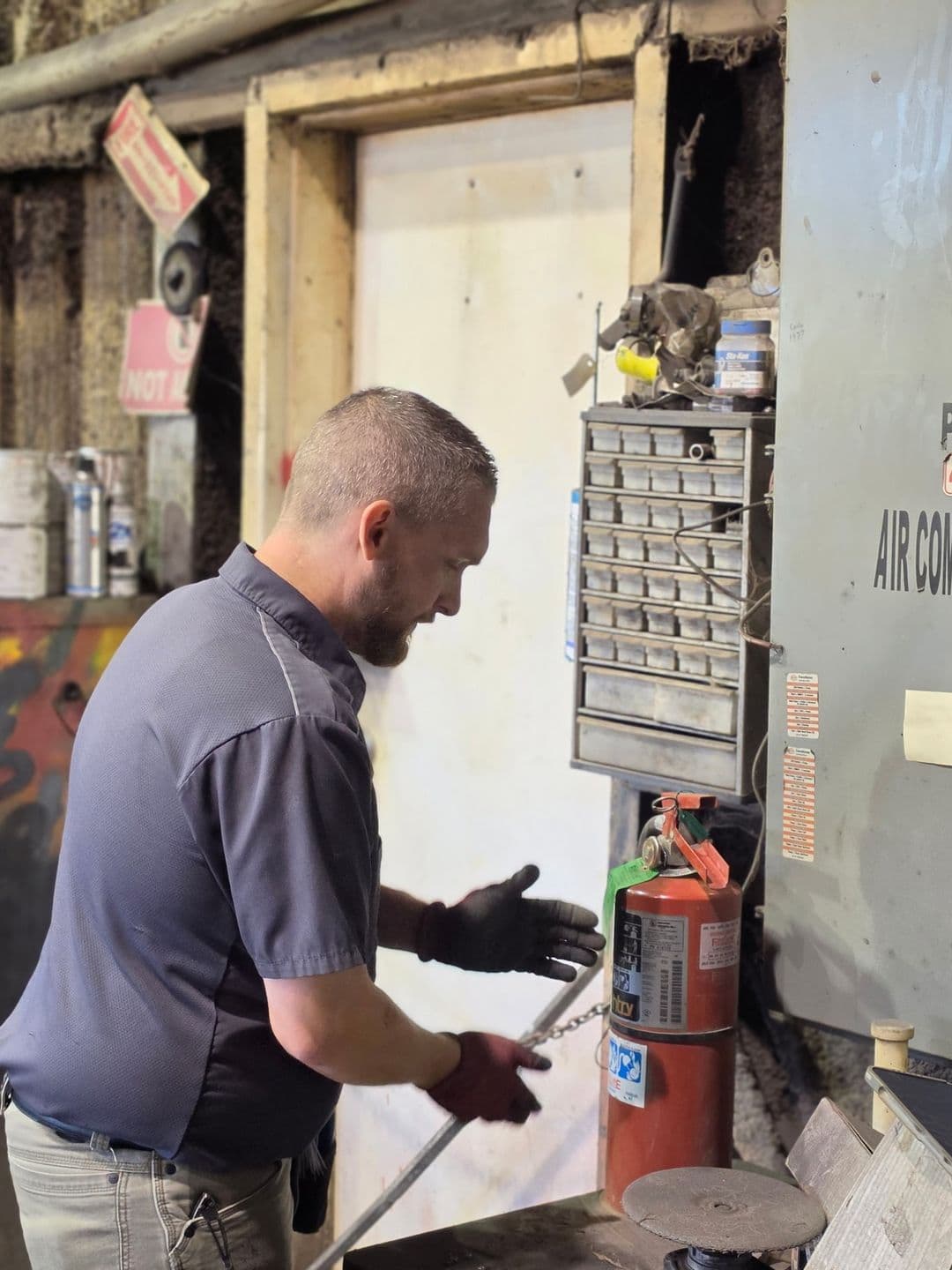 First-Line technician inspecting a fire extinguisher beside an air compressor panel in an industrial shop