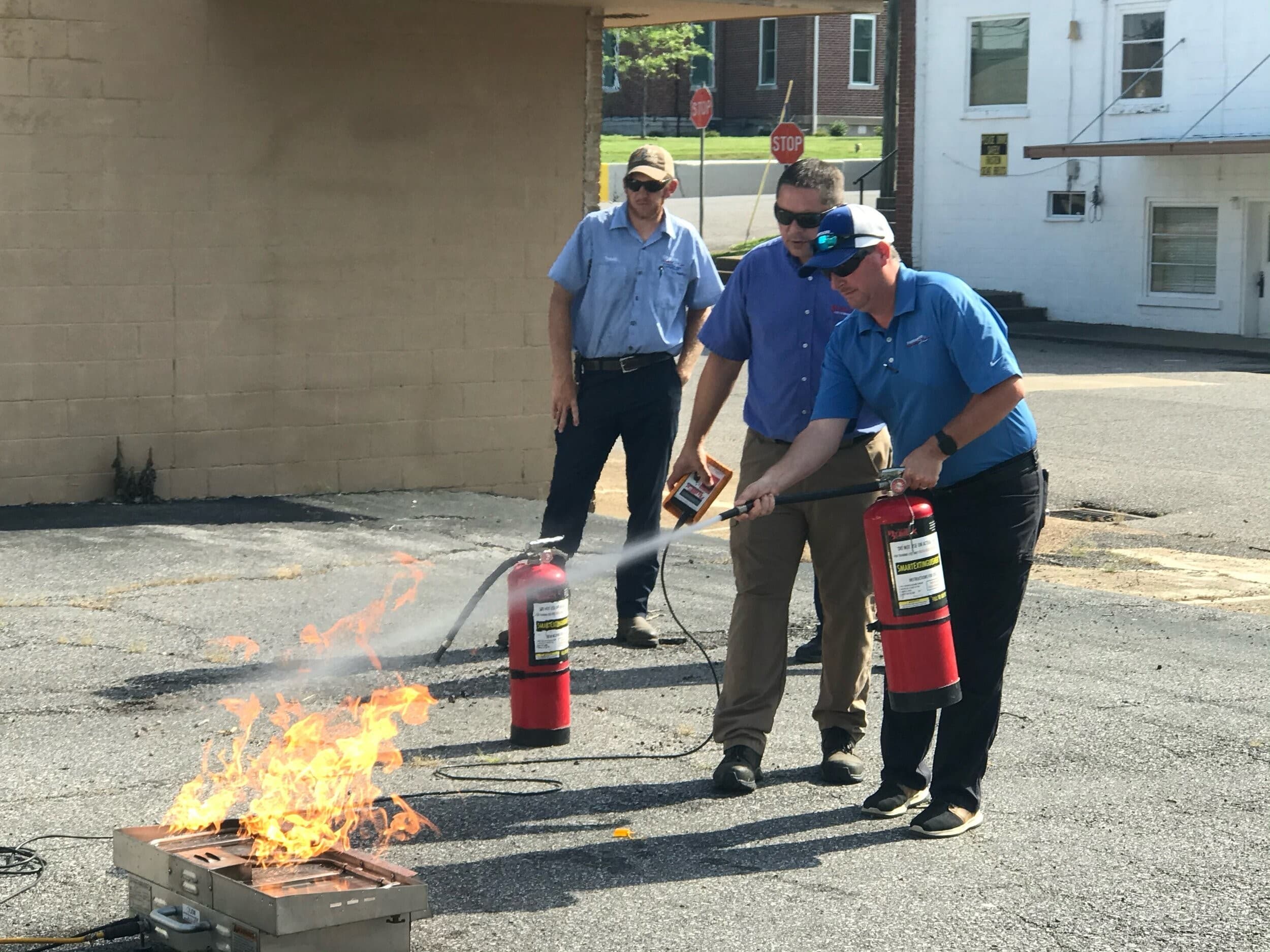 First-Line techs on a live-fire training: one discharging an extinguisher at a fire pan, instructor with the burn-tray remote