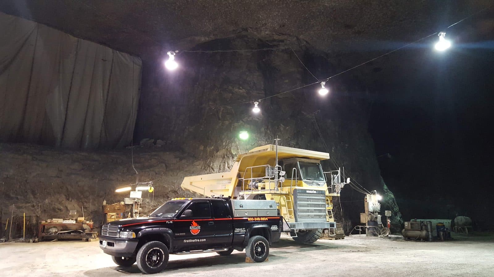 A First-Line service pickup parked beside a yellow haul truck in an underground mine, lit by hanging work lights